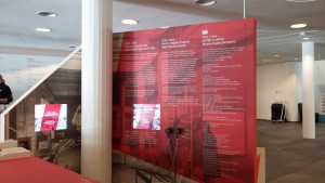 Ausstellungsstation im Foyer des DSM. Bildschirme mit Dokumentarfilmen, in der Mitte Hörmuscheln, dreisprachige Textwand.